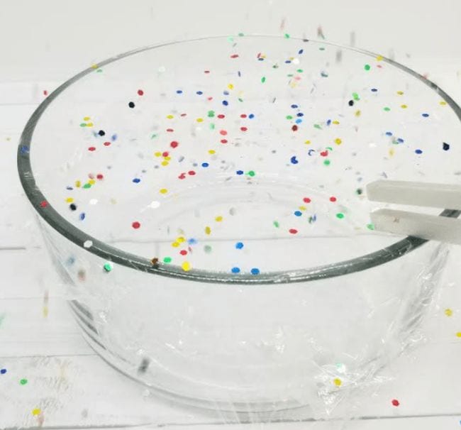 Glass bowl covered in plastic wrap with confetti on top, and a tuning fork touching the plastic
