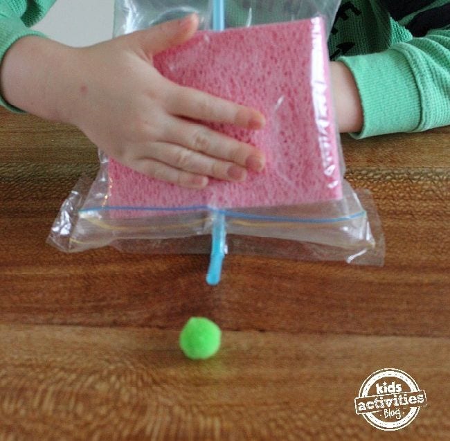 Young student using a straw in a plastic bag with sponges to move a green pompom across a wood table